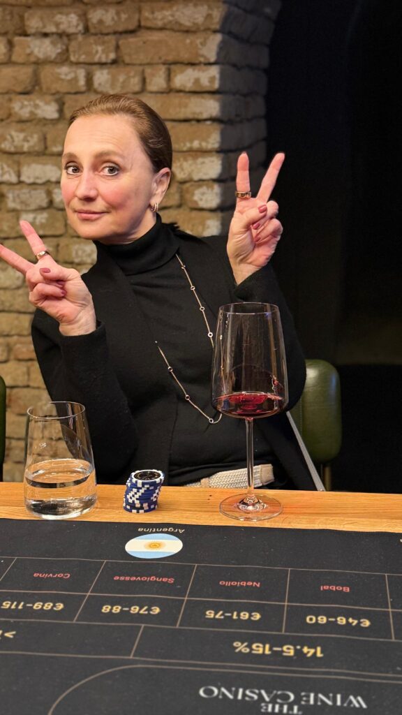 Eine Frau sitzt an einem Casinotisch mit Pokerchips, einem Glas Rotwein und Wasser vor sich, lächelt und blinkt mit zwei Peace-Zeichen. Es ist die perfekte Szene für einen Wein-Brettspiel-Abend vor einer gemütlichen Backsteinmauer-Kulisse.