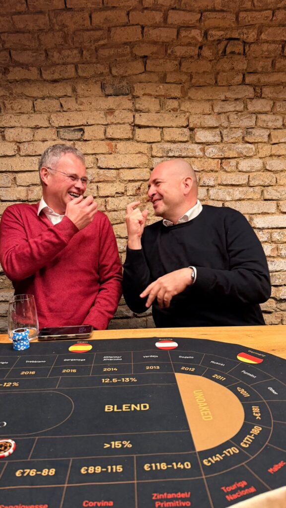Zwei Männer sitzen an einem Tisch mit einer Spielmatte, lachen und heben die Hände, als würden sie auf einen Wein-Brettspiel-Abend anstoßen. Der eine trägt einen roten Pullover, der andere einen schwarzen. Beide sind von einer rustikalen Backsteinwand eingerahmt und genießen die spielerische Atmosphäre.