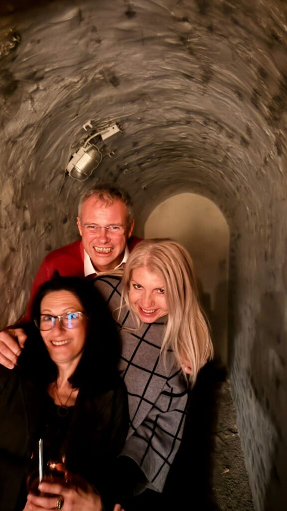 Drei Erwachsene, zwei Frauen und ein Mann, lächeln gemeinsam in einem engen, schwach beleuchteten Tunnel mit rauen Steinwänden - vielleicht sind sie auf dem Weg zu einem Wein-Brettspiel-Abend. Warmes Licht beleuchtet ihre Gesichter, während sich der Tunnel in den Hintergrund erstreckt.