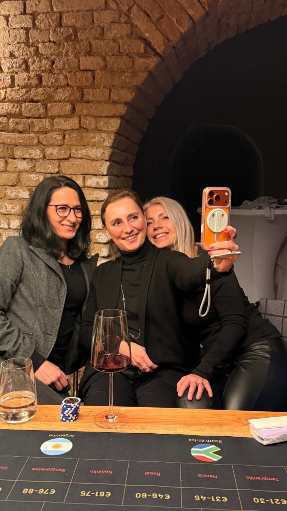 Drei Frauen sitzen lächelnd beieinander und posieren für ein Selfie bei einem gemütlichen Wein-Brettspiel-Abend, mit Weingläsern und Pokerchips auf dem Tisch in einem gemauerten Raum. Eine Frau hält ihr Handy hoch, um den lustigen Moment festzuhalten.