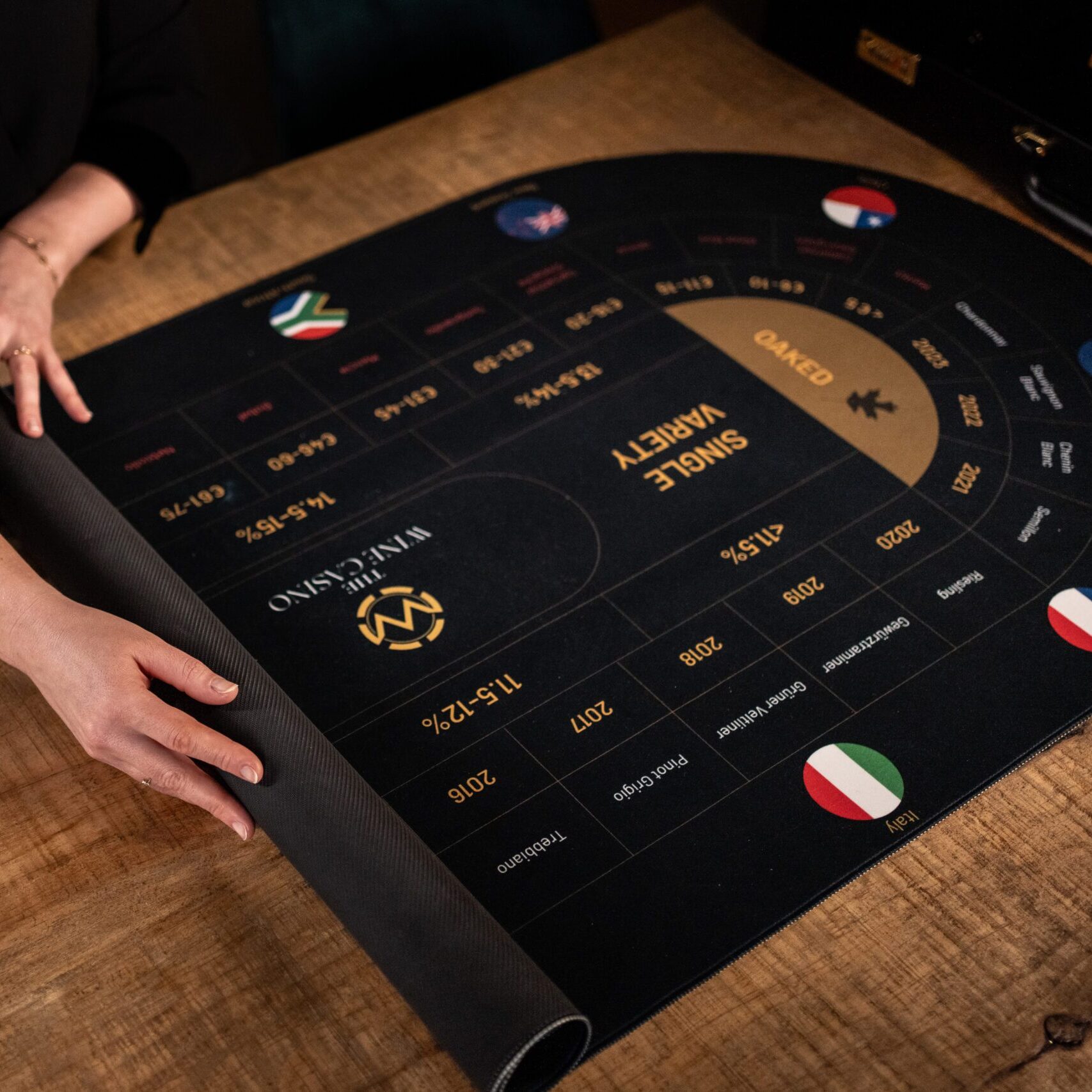 Eine Person rollt eine schwarze Casinospielmatte mit goldenem Text und verschiedenen Flaggen auf einem Holztisch aus. Die Matte zeigt Wettoptionen und Quoten in Abschnitten für verschiedene Spielarten an.