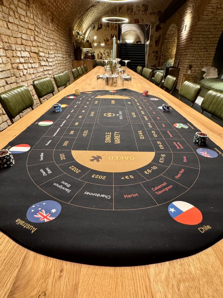 Ein langer Holztisch in einem gemauerten Raum ist wie ein Casinotisch aufgebaut, perfekt für einen Wein-Brettspiel-Abend, mit Chips und einer schwarzen Filzmatte, auf der Ländernamen, Jahreszahlen und Routen stehen. Grüne Stühle stehen auf beiden Seiten des Tisches.