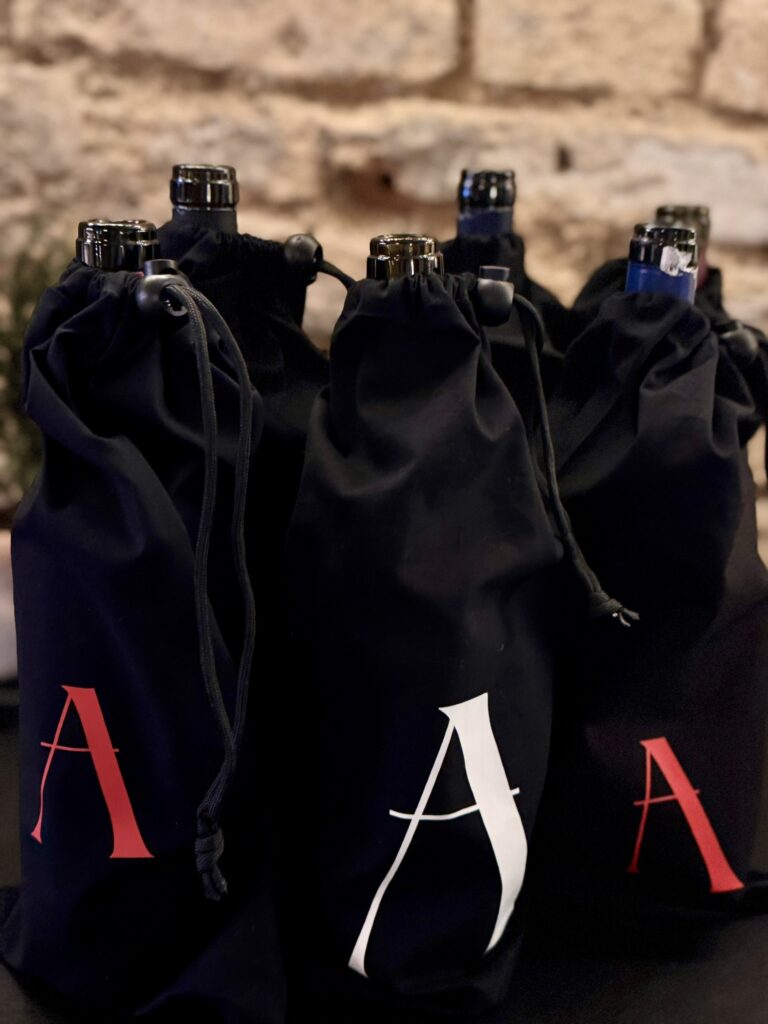 Vor einem verschwommenen Steinmauer-Hintergrund stehen mehrere Weinflaschen in schwarzen Kordeltaschen, jede mit einem großen, stilisierten Buchstaben A in Rot oder Weiß - perfekt für einen spannenden Wein-Brettspiel-Abend.