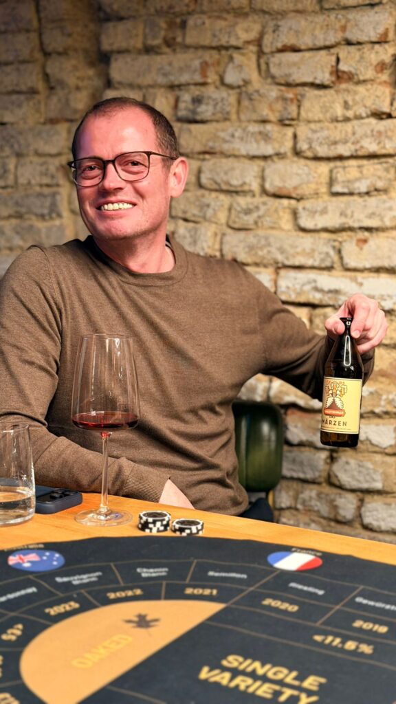 Eine lächelnde Person mit Brille und braunem Pullover sitzt an einem Tisch und genießt einen Wein-Brettspiel-Abend mit einem Weinglas, einer Apfelweinflasche, Pokerchips und einem Spielbrett mit Ländern und Jahreszahlen. Im Hintergrund ist eine Backsteinmauer zu sehen.