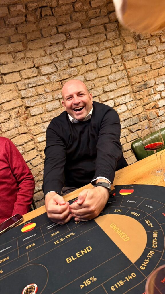 Ein lächelnder Mann in einem schwarzen Pullover sitzt an einem Tisch mit einem Weinglas und einer Probiermatte und streckt spielerisch beide Hände nach vorne - bereit für einen unterhaltsamen Wein-Brettspiel-Abend. Im Hintergrund ist eine rustikale Backsteinmauer zu sehen.