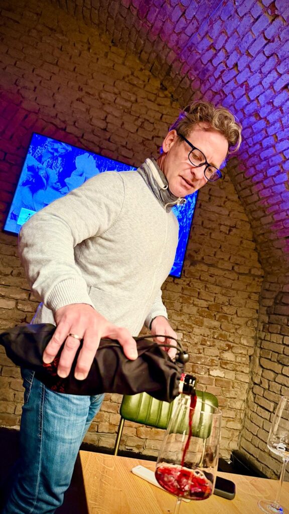 Ein Mann mit Brille und hellem Pullover schenkt bei einem Wein-Brettspiel-Abend Rotwein aus einer in schwarzes Tuch gewickelten Flasche ein. Er steht in einem gemauerten Raum mit gewölbter Decke, einem Stuhl und einem Fernsehbildschirm im Hintergrund.