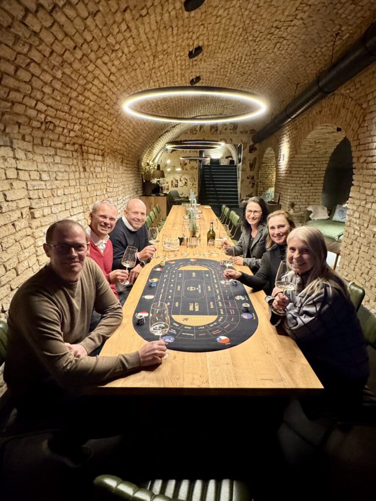 Sechs Personen sitzen um einen langen Holztisch in einem Raum mit Backsteinbögen und genießen einen warmen und gemütlichen Wein-Brettspiel-Abend. Lächelnd und mit Weingläsern in der Hand spielen sie ein Brettspiel, auf dem Tisch stehen mehrere Flaschen, über dem Tisch herrscht gedämpftes Licht.